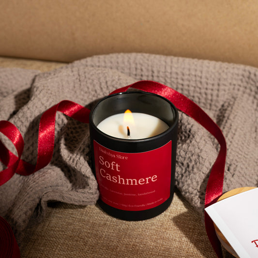 A lit candle in a amber jar with a red label reading  Soft Cashmere,' styled on a beige fabric surface with a gray knit blanket and a red ribbon.