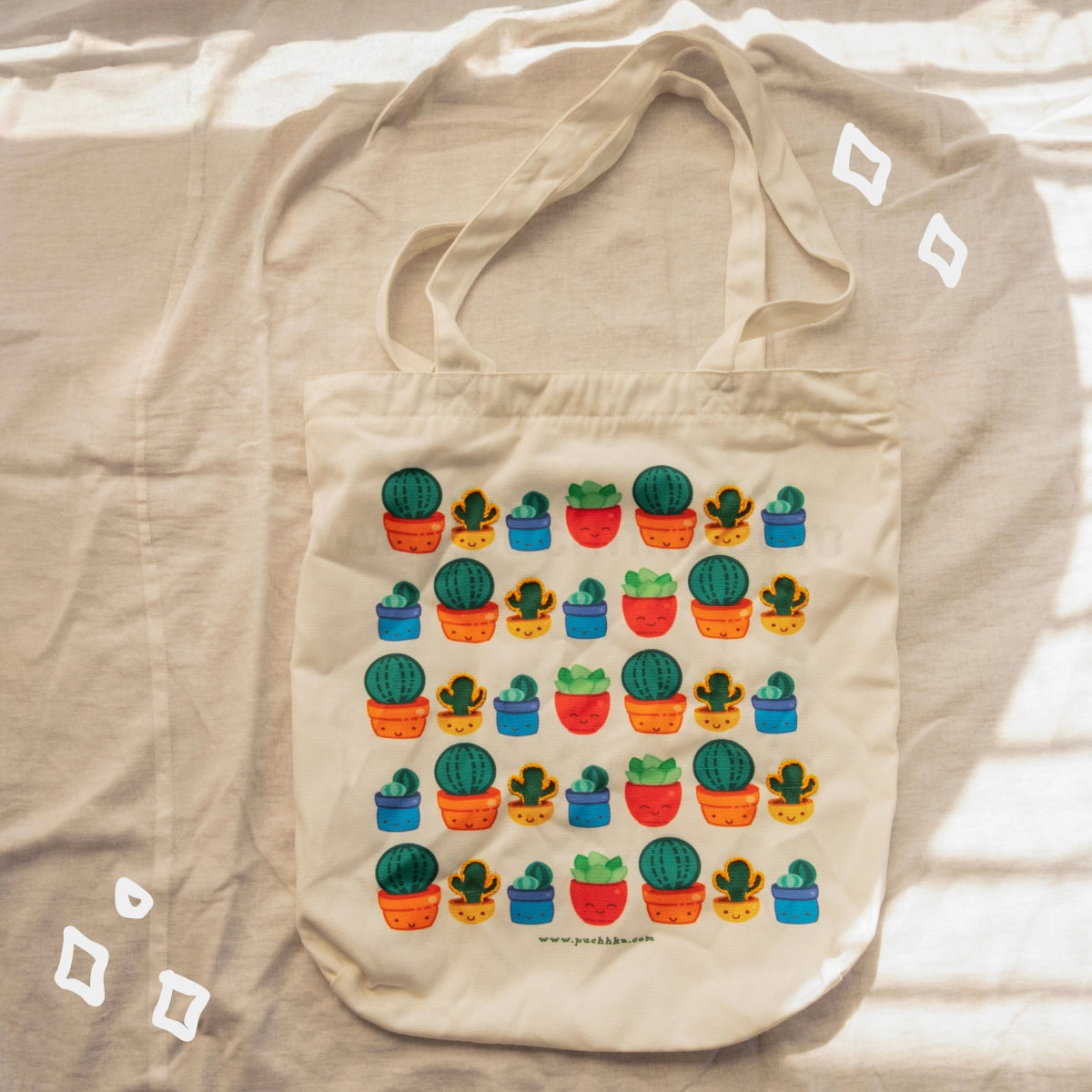 A beige canvas tote bag with a colorful cactus and succulent pattern, laid flat on a beige fabric background with soft shadows.