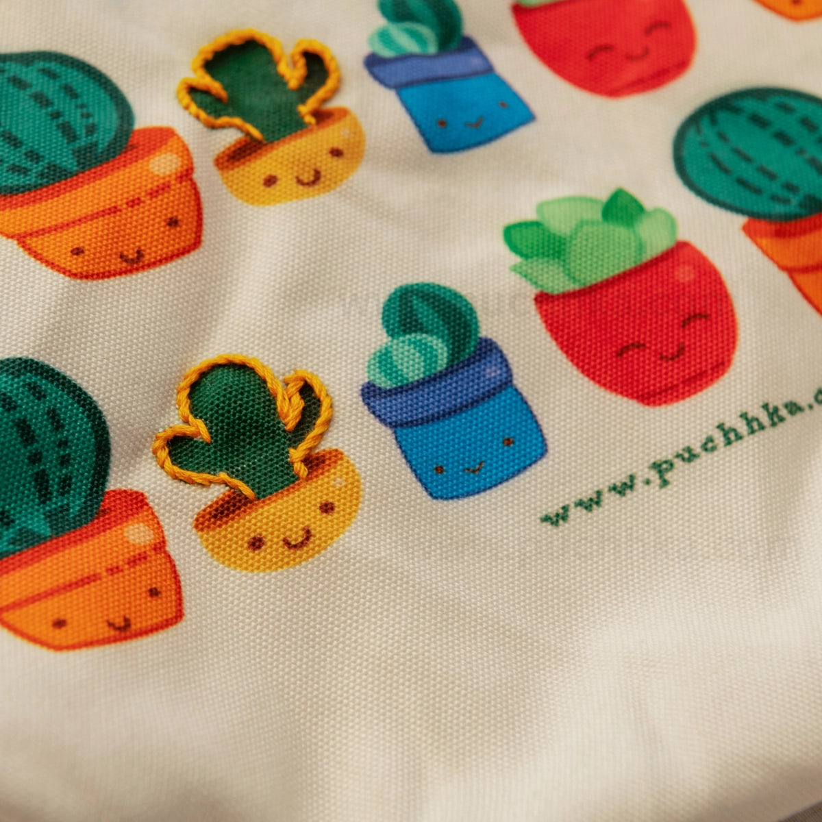 Close-up of a cream-colored fabric surface printed and hand embroidered with colorful potted cacti and succulents.