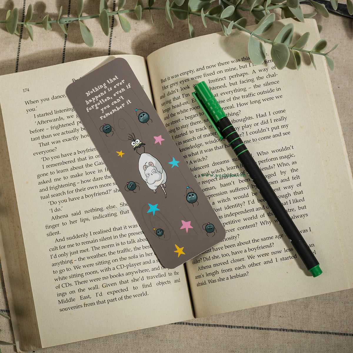 A rectangular book bookmark with a ghibli character and colorful stars, placed on an open book next to a green pen.