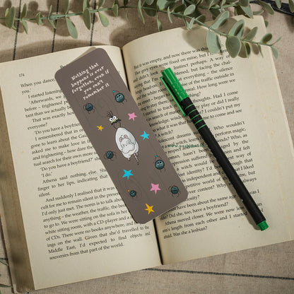 A rectangular book bookmark with a ghibli character and colorful stars, placed on an open book next to a green pen.