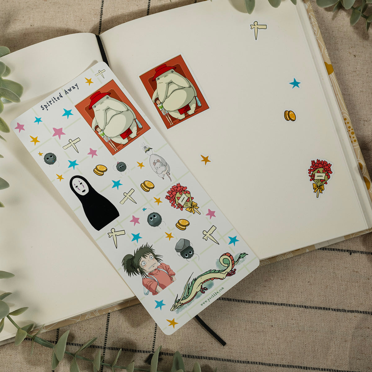 A decorative sticker sheet with anime and pop culture illustrations, including No-Face from Spirited Away, is placed on an open notebook. The sticker sheet features a white base with colorful cartoon characters, stars, and symbols, styled on a neutral fabric background with green leaves.