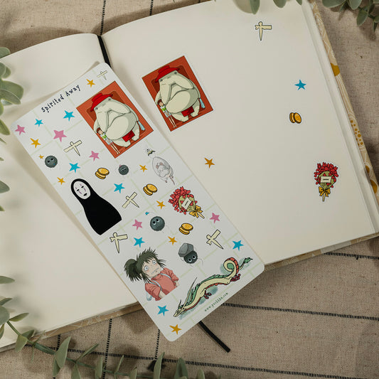 A decorative sticker sheet with anime and pop culture illustrations, including No-Face from Spirited Away, is placed on an open notebook. The sticker sheet features a white base with colorful cartoon characters, stars, and symbols, styled on a neutral fabric background with green leaves.