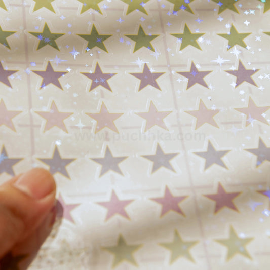 A close-up of a sheet of star-shaped stickers in muted green, pink, and purple tones with a subtle holographic shimmer, held by a finger in the lower left corner.