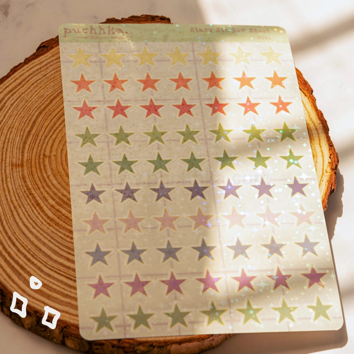 A sheet of decorative star stickers in multiple pastel colors, including yellow, orange, green, pink, purple, and blue, arranged in rows on a translucent backing. The sheet is placed on a round wooden coaster with soft sunlight and shadows across it.