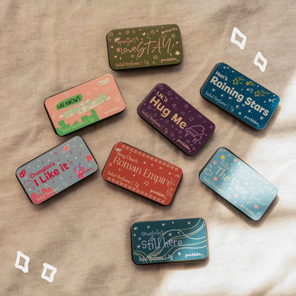 Eight small rectangular solid perfume tin boxes arranged on a beige fabric background, each with a different illustrated design and inspired by members of Stray Kids.