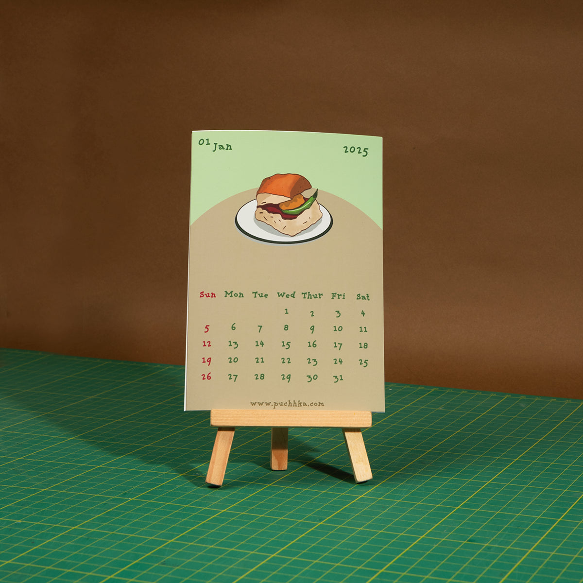 A novelty monthly calendar card featuring a hand-drawn hamburger illustration, displayed on a small wooden easel stand over a green cutting mat against a brown backdrop.