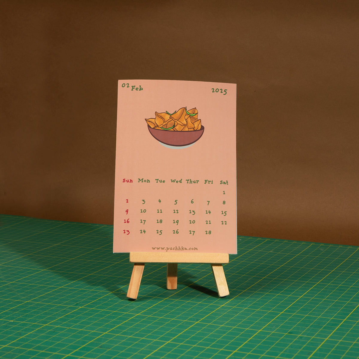 A miniature paper calendar page from February 2025 stands on a small wooden easel. The page is pale pink with a small illustration of a bowl of chips and the date 03 Feb at the top.