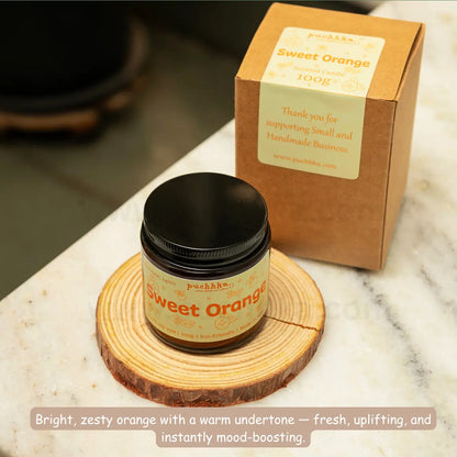 A jar of Puchhka Sweet Orange scented candle placed on a round wooden coaster, with its kraft paper box standing behind it on a white marble surface.