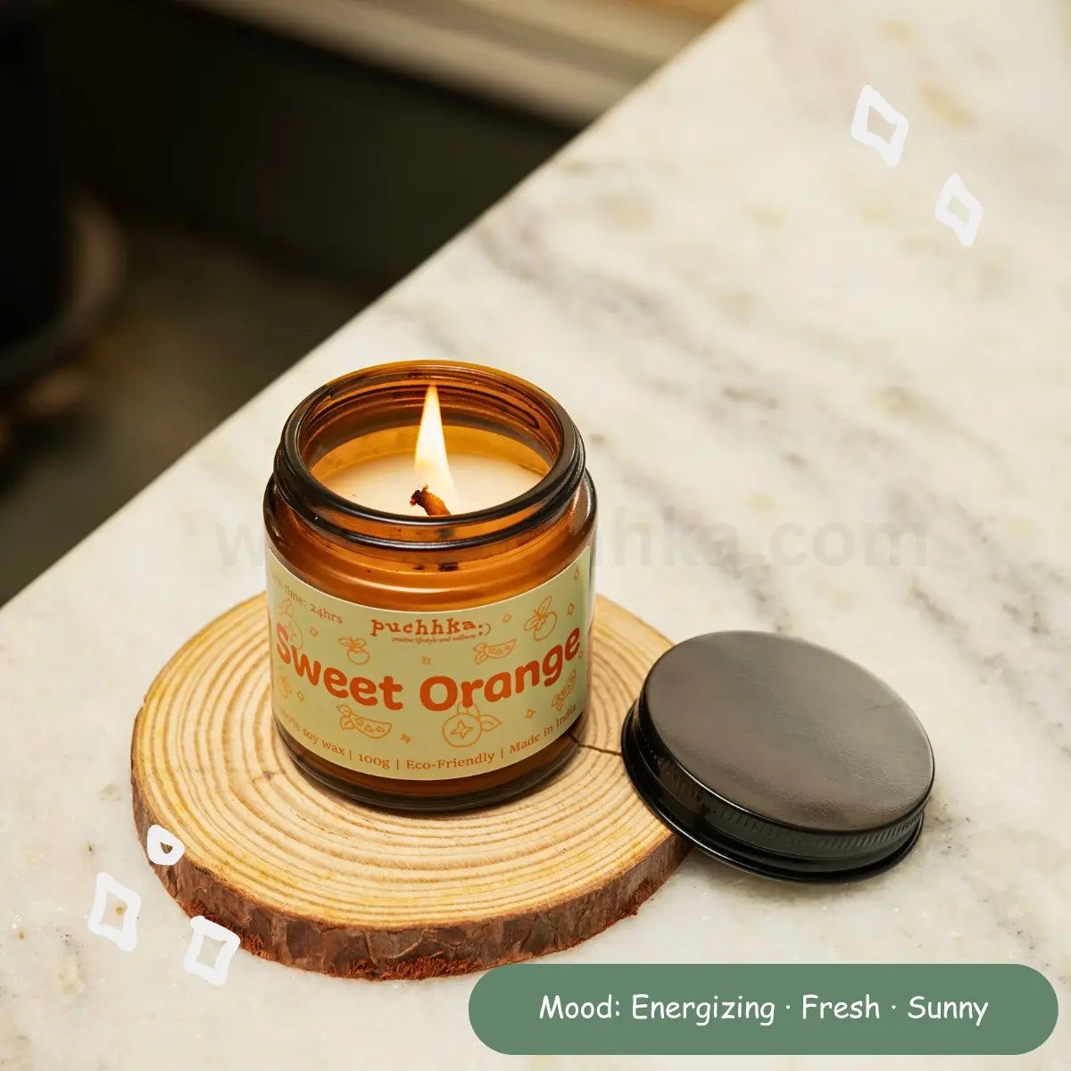 An amber glass jar candle labeled 'puchhka Sweet Orange' is lit on a round wood  coaster, with its black lid placed beside it on a white marble surface. A green text bubble at the bottom reads 'Mood: Energizing • Fresh • Sunny'.