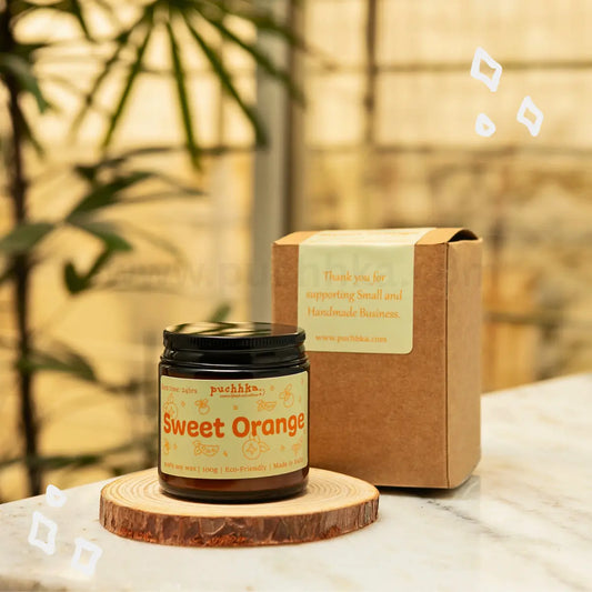 A jar of Puchhka "Sweet Orange" scented candle with a black lid, displayed on a wooden coaster next to its brown kraft gift box on a marble surface.