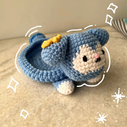 A handmade crocheted Tasy plush Tray with cream-colored face and paws, a small yellow bow, and black embroidered eyes, resting on a beige stone surface.
