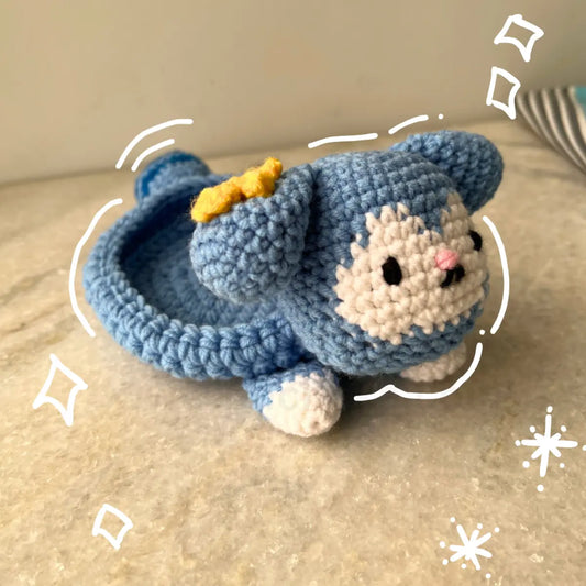A handmade crocheted Tasy plush Tray with cream-colored face and paws, a small yellow bow, and black embroidered eyes, resting on a beige stone surface.