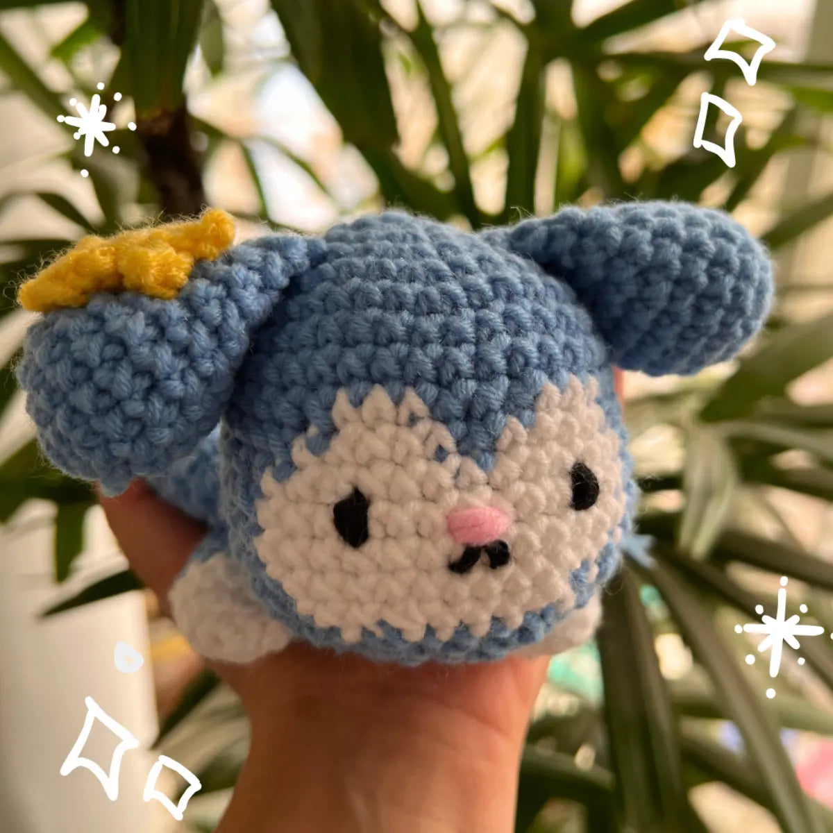 A handmade crocheted Tasy Plush Tray held in a hand, featuring a yellow bow on its left ear, black embroidered eyes and mouth, and a small pink nose, with blurred green leaves in the background.