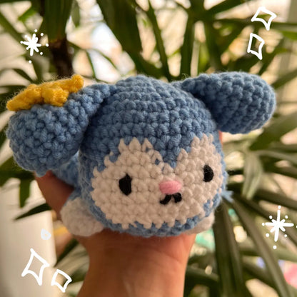 A handmade crocheted Tasy Plush Tray held in a hand, featuring a yellow bow on its left ear, black embroidered eyes and mouth, and a small pink nose, with blurred green leaves in the background.