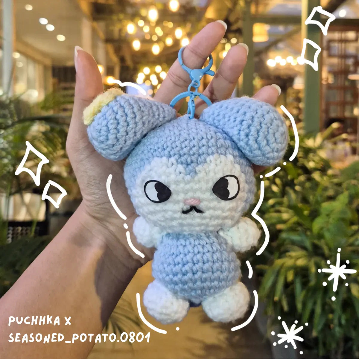 A handmade crocheted Tasy keychain held in a hand, featuring light blue and white yarn with embroidered facial details, set against a blurred background of a shop interior with warm lights and greenery.