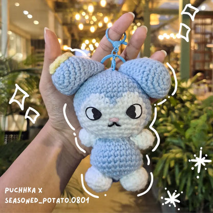 A handmade crocheted Tasy keychain held in a hand, featuring light blue and white yarn with embroidered facial details, set against a blurred background of a shop interior with warm lights and greenery.