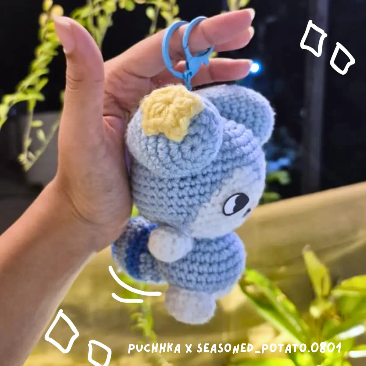 A hand holds a crocheted Tasy keychain with a yellow star on its head and a blue key ring, against a dark background with blurred green plants.