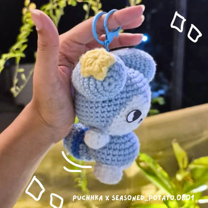 A hand holds a crocheted Tasy keychain with a yellow star on its head and a blue key ring, against a dark background with blurred green plants.