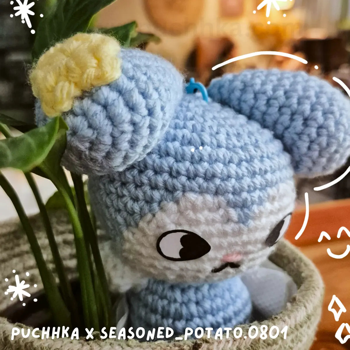 A crocheted Tasy plush Keychain with a light blue body, white face area, and a yellow bow on its head, posed among green plant leaves in a basket. The image has decorative white doodle overlays and text reading 'PUCHHKA X SEASONED_POTATO_0801' at the bottom.
