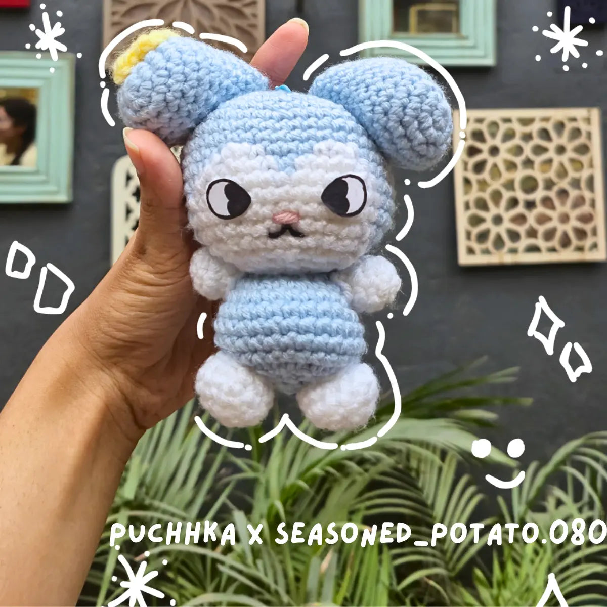 A handmade crocheted Tasy held in a person's hand, featuring light blue and white yarn with black embroidered eyes and a small pink nose. The background shows a dark wall with framed pictures and green plants.