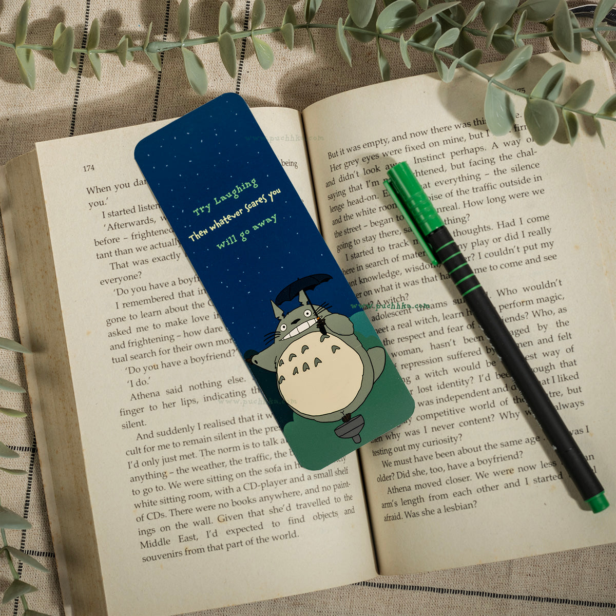 A small Totoro character bookmark , placed on an open book with a green pen resting on top.