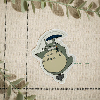 A die-cut vinyl sticker of the character Totoro from Totoro! on a textured beige linen background with decorative green leaves.
