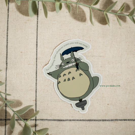 A die-cut vinyl sticker of the character Totoro from Totoro! on a textured beige linen background with decorative green leaves.