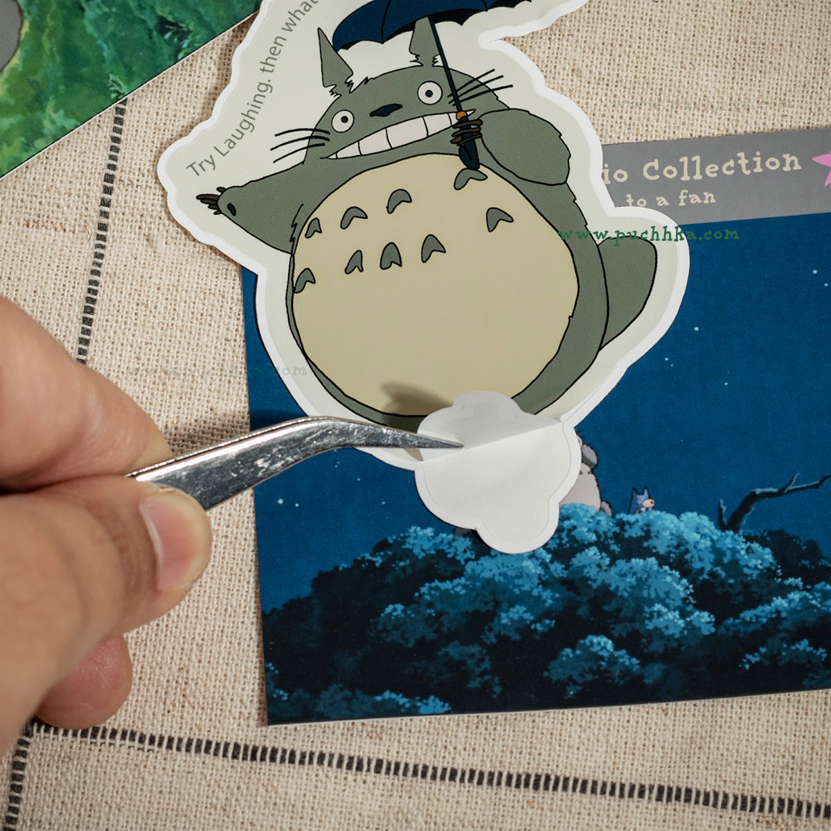 A hand using tweezers to peel a white cloud-shaped sticker from a Totoro die-cut sticker, placed on top of a printed card over a beige woven fabric background.