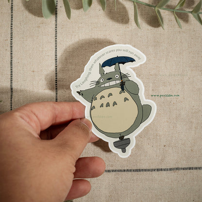 A hand holds a die-cut sticker of Totoro from My Neighbor Totoro against a beige linen background with green leaves at the top.