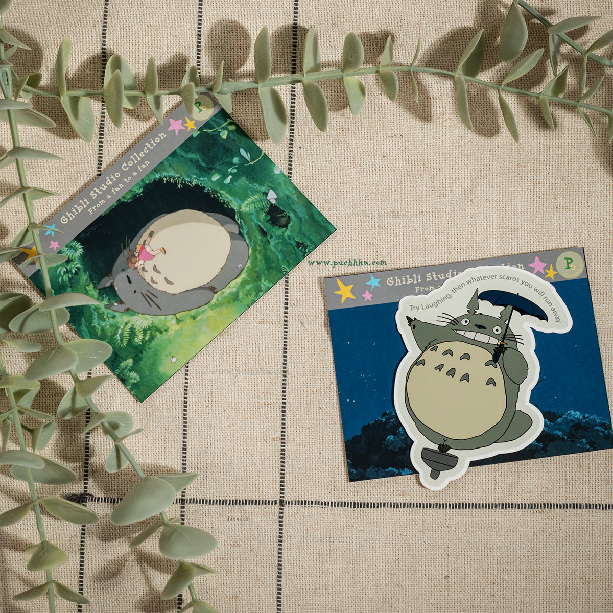 Two Totoro-themed sticker cards displayed on a beige linen background with eucalyptus leaves. One card shows Totoro in a green forest, and the other shows Totoro with a bow on a dark blue night background.