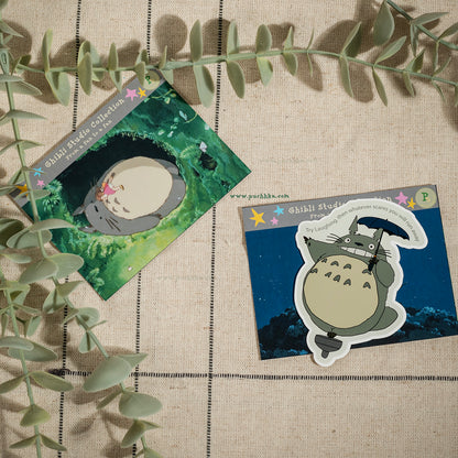 Two Totoro-themed sticker cards displayed on a beige linen background with eucalyptus leaves. One card shows Totoro in a green forest, and the other shows Totoro with a bow on a dark blue night background.