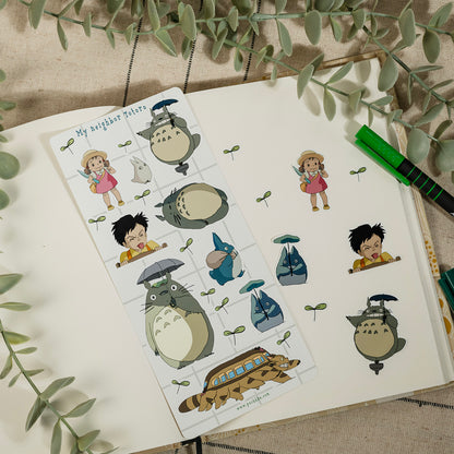 A sheet of Totoro-themed planner stickers displayed on an open notebook, surrounded by decorative green leaves and a green pen.