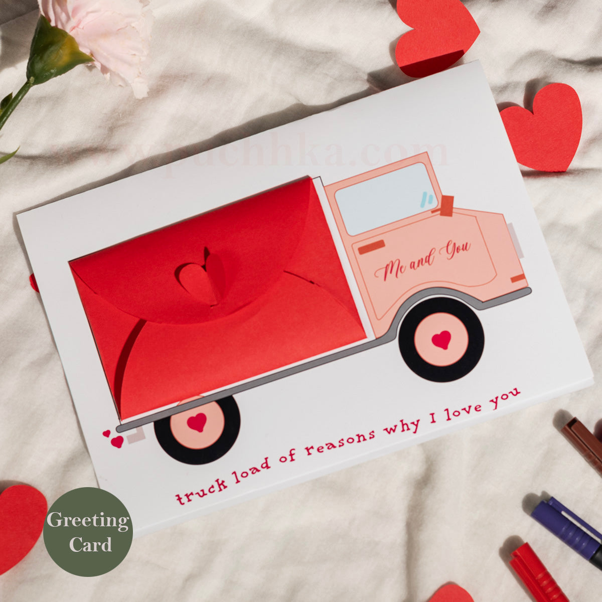 A Valentine's Day greeting card featuring a  red truck with the words 'Me and You' and 'truck load of reasons why I love you'. The card is displayed on a textured white fabric with red heart cutouts and a pink flower.