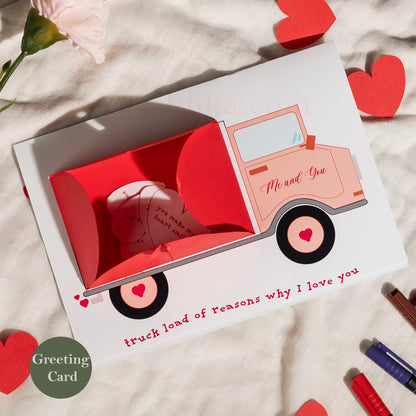 A pop-up greeting card featuring a pink truck carrying folded paper hearts, styled on a textured cream fabric with scattered red hearts and a pink flower.