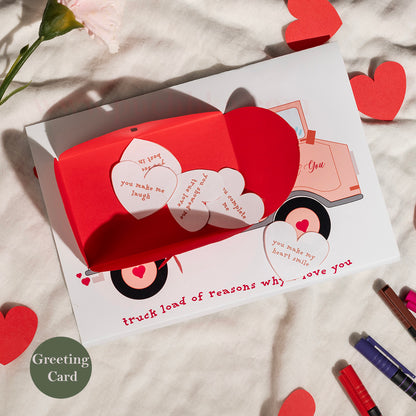 A folded white greeting card with a red pop-up truck and heart-shaped message pieces, styled on a textured white fabric with red heart cutouts and a flower stem.