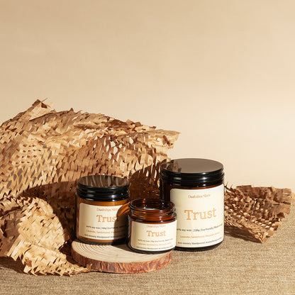 Three Daakshya Store (now puchhka) soy wax candles in amber glass jars with black lids and cream labels, styled on a wood slice with crumpled paper against a beige background.