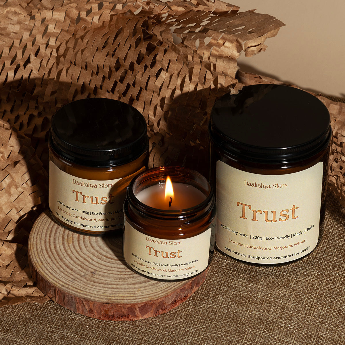 Three jars of Daakshya Store's  'Trust' soy wax candles arranged on a rustic wood coaster and woven brown paper backdrop. One candle is lit in the center, flanked by two sealed amber glass jars with cream labels and black lids.