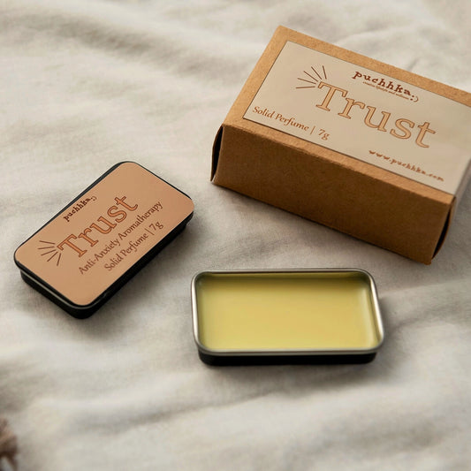 A small rectangular metal tin of solid perfume balm sits open on a light fabric surface, with its kraft paper box and a second closed tin nearby. The labels read 'puchhka' and 'Trust'.