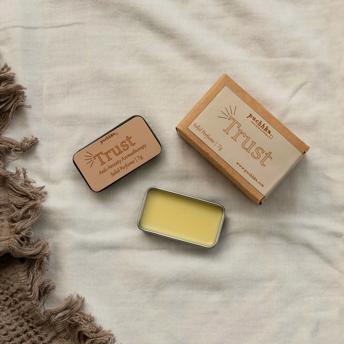 A styled flat-lay of a solid perfume product on a textured beige fabric background. The set includes a kraft paper box labeled 'trust' by 'puchhka', a small rectangular tin with a tan label, and an open silver tin showing pale yellow wax.