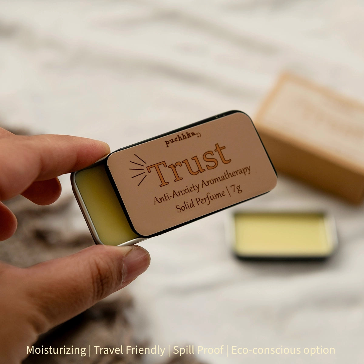 A hand holds a small rectangular metal tin of solid perfume labeled 'Trust' by Puchhka. The tin shows pale yellow wax and a tan label with dark brown text.