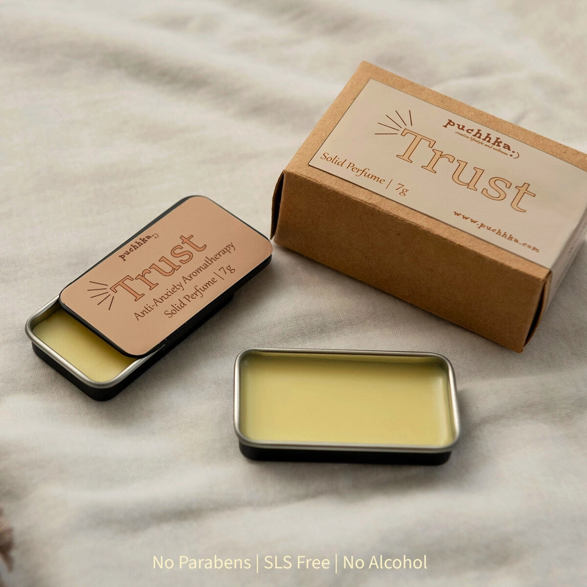 An open metal tin of Puchhka's Trust solid perfume balm next to its kraft paper packaging box, with a second closed tin showing the lid label. The balm is a pale yellow color, set on a textured beige fabric background.