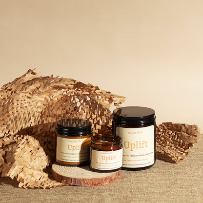 Three amber glass jars with cream labels and black lids, labeled 'Uplift', arranged on a wood  with decorative woven paper against a warm beige background.