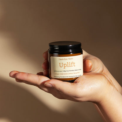 A hand holding a small amber jar candle labeled 'Daakshya Store Uplift' against a warm beige background.