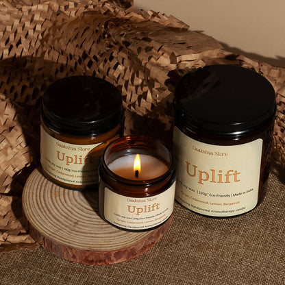  'Uplift' candles by Daakshya Store are arranged on a rustic wood coaster. One candle is lit in a small amber glass jar in the center, while two larger unlit amber glass jars with black lids sit on either side.