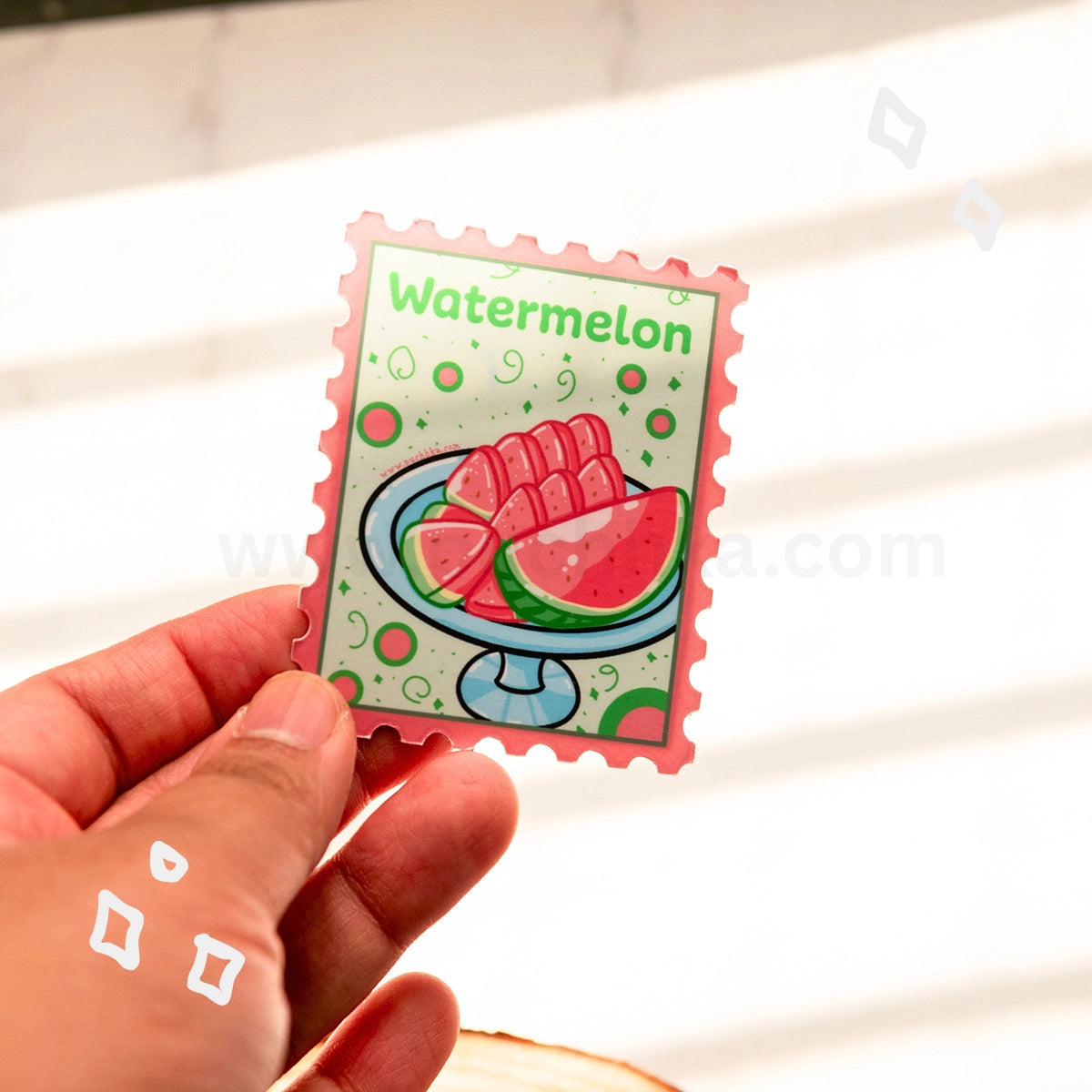 A hand holds a small rectangular magnet with a watermelon-themed illustration and the word "Watermelon" at the top. The design shows watermelon slices on a pedestal plate with green and pink details, photographed against a bright white background with soft shadows.