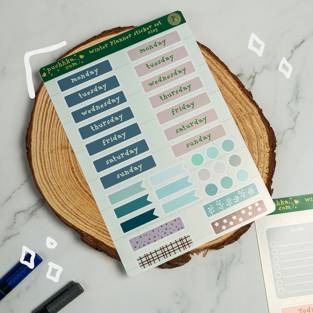 A sheet of planner sticker accessories displayed on a round wood slice coaster over a white marble surface. The sticker sheet includes pastel blue weekday labels, pink weekday labels, light blue and teal circles, lavender patterned strips, and small decorative accents.